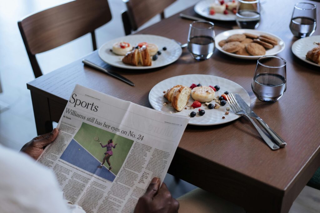 A man reads the sports section while enjoying breakfast at a neatly set table indoors.